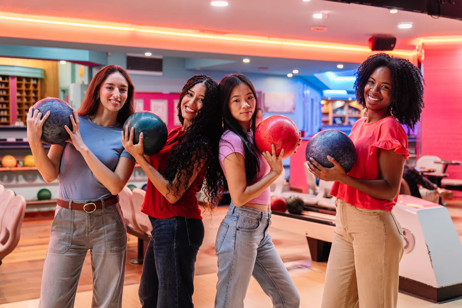 Vier junge Frauen mit unterschiedlichen Gesichtern halten Bowlingkugeln in der Hand und genießen lächelnd ein freundschaftliches Spiel auf der Bowlingbahn.